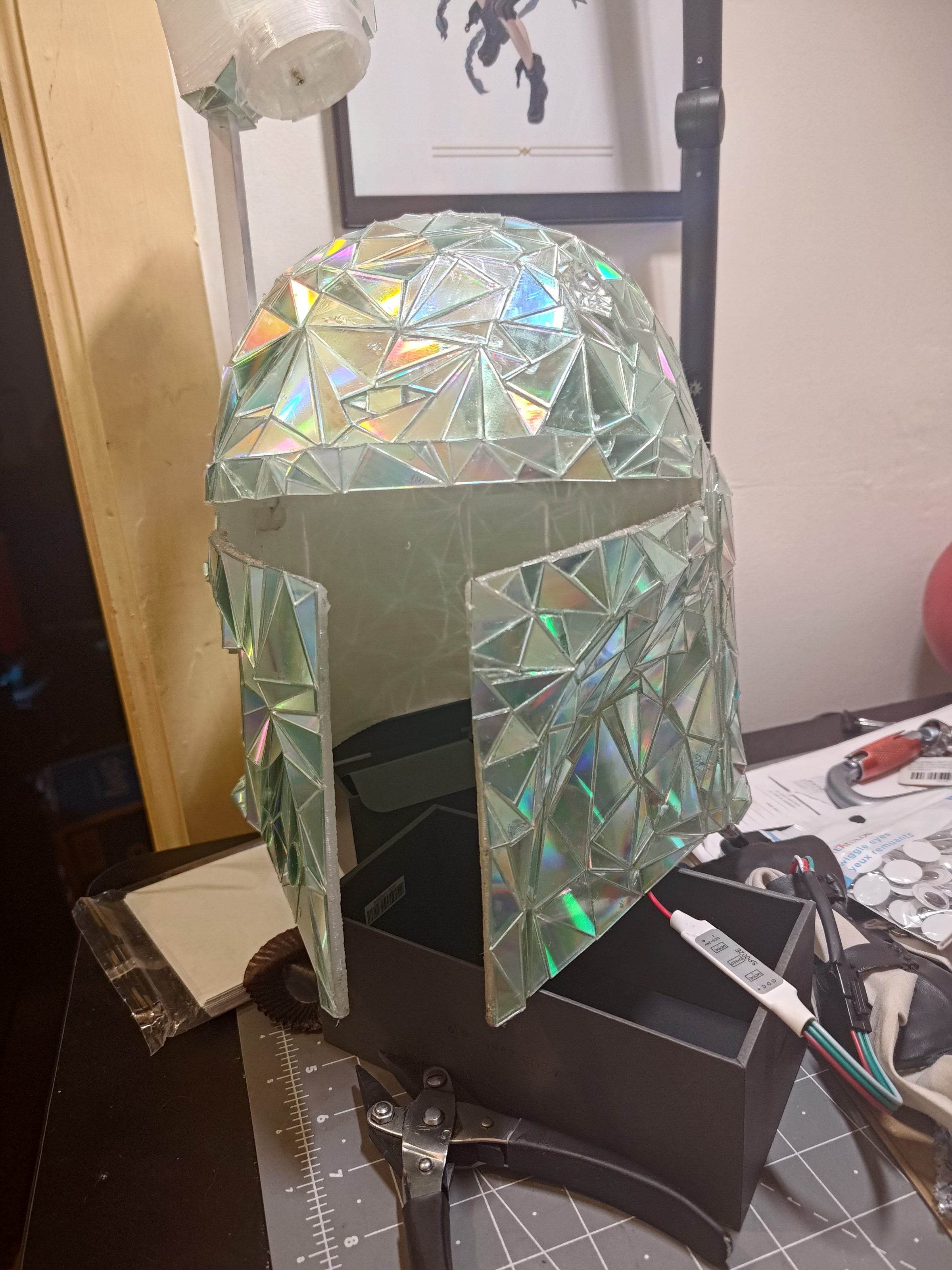 A Mandalorian-style helmet tiled in iridescent dichroic film, on a stand in the workshop.