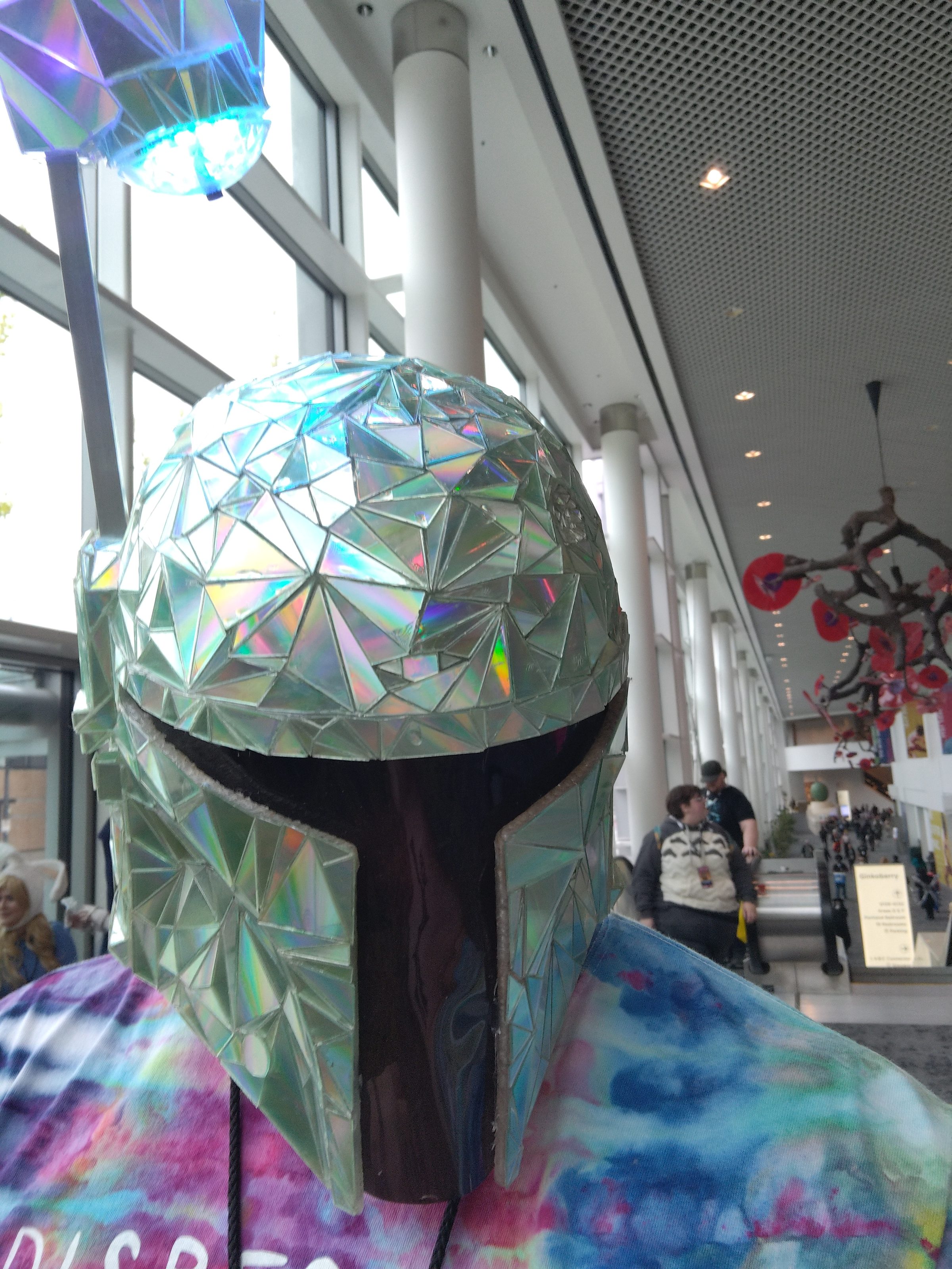 The dichroic-tiled Mandalorian helmet worn at a convention, its facets catching the daylight.