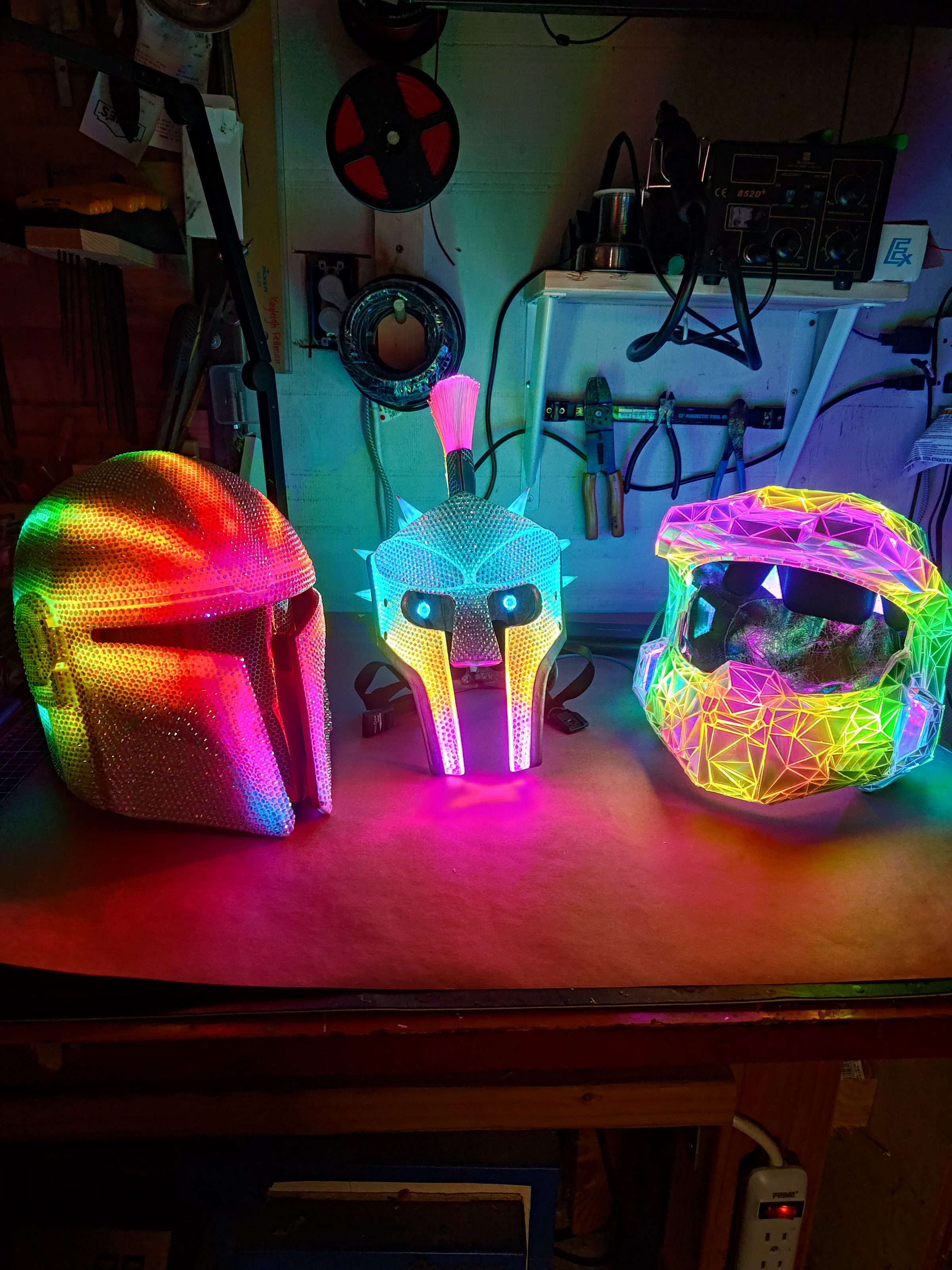 Three illuminated helmets lined up on a workbench, each lit a different colour.