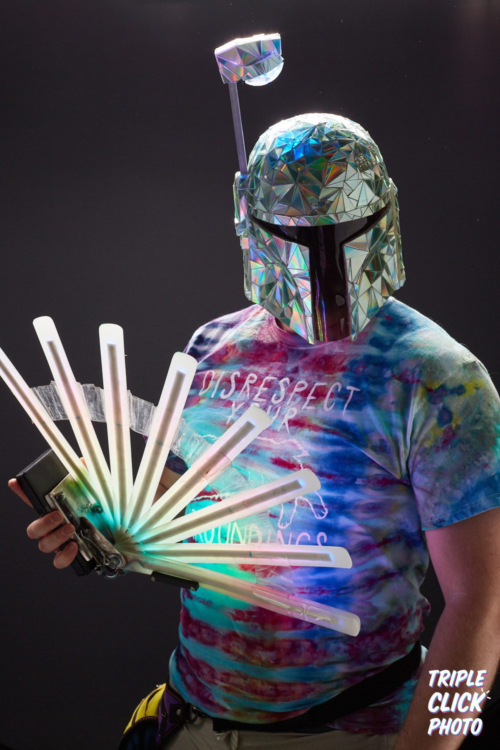 Studio portrait of the dichroic-tiled Mandalorian helmet, a fanned rack of glowing light tubes held across the chest.