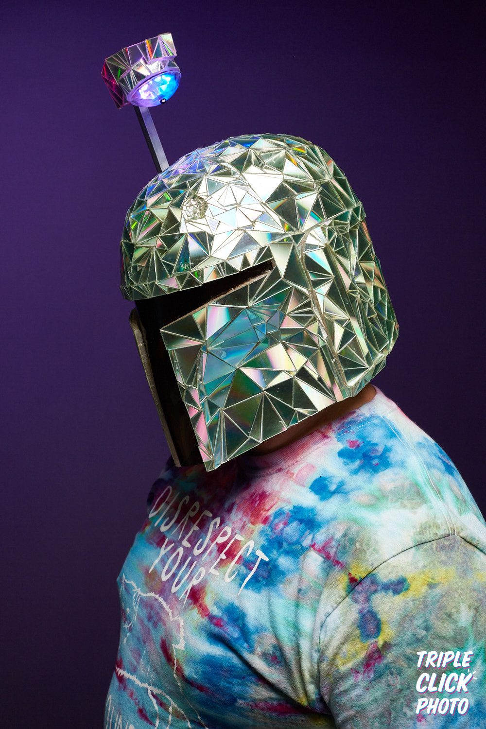 The dichroic-tiled Mandalorian helmet in profile against purple, the small lamp on its antenna lit blue.