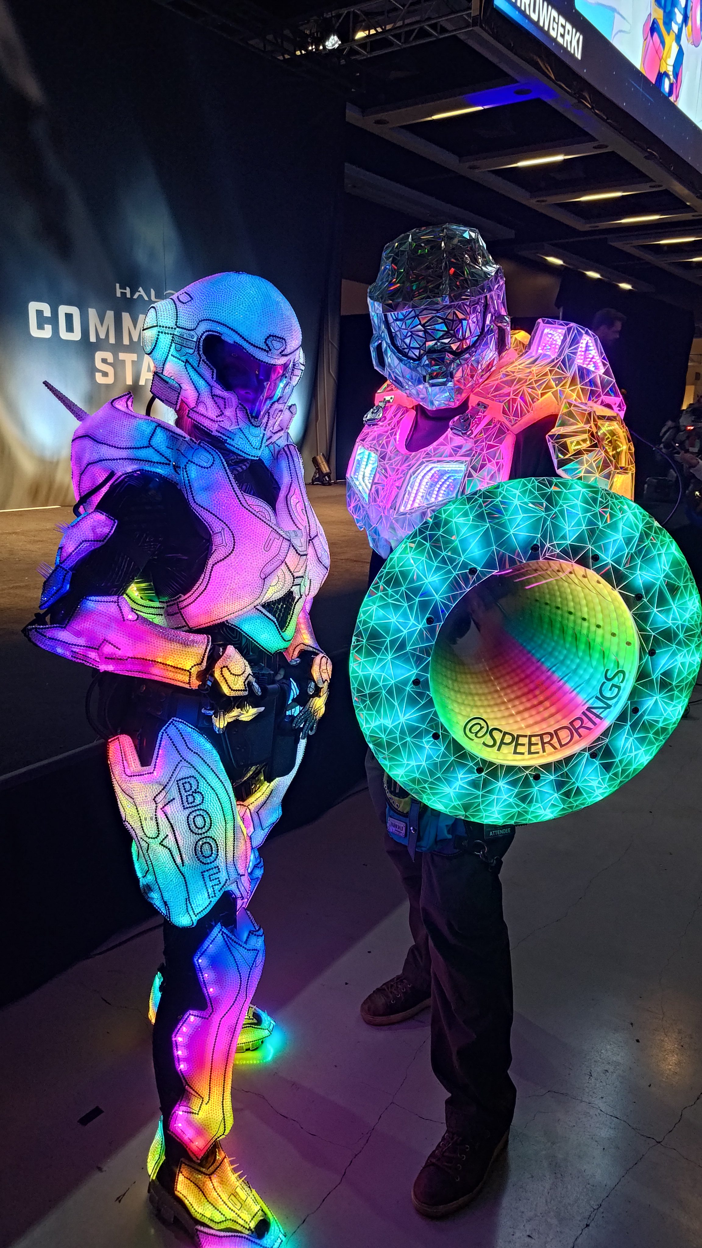 Two armoured cosplayers at a Halo convention booth, one holding the illuminated shield.