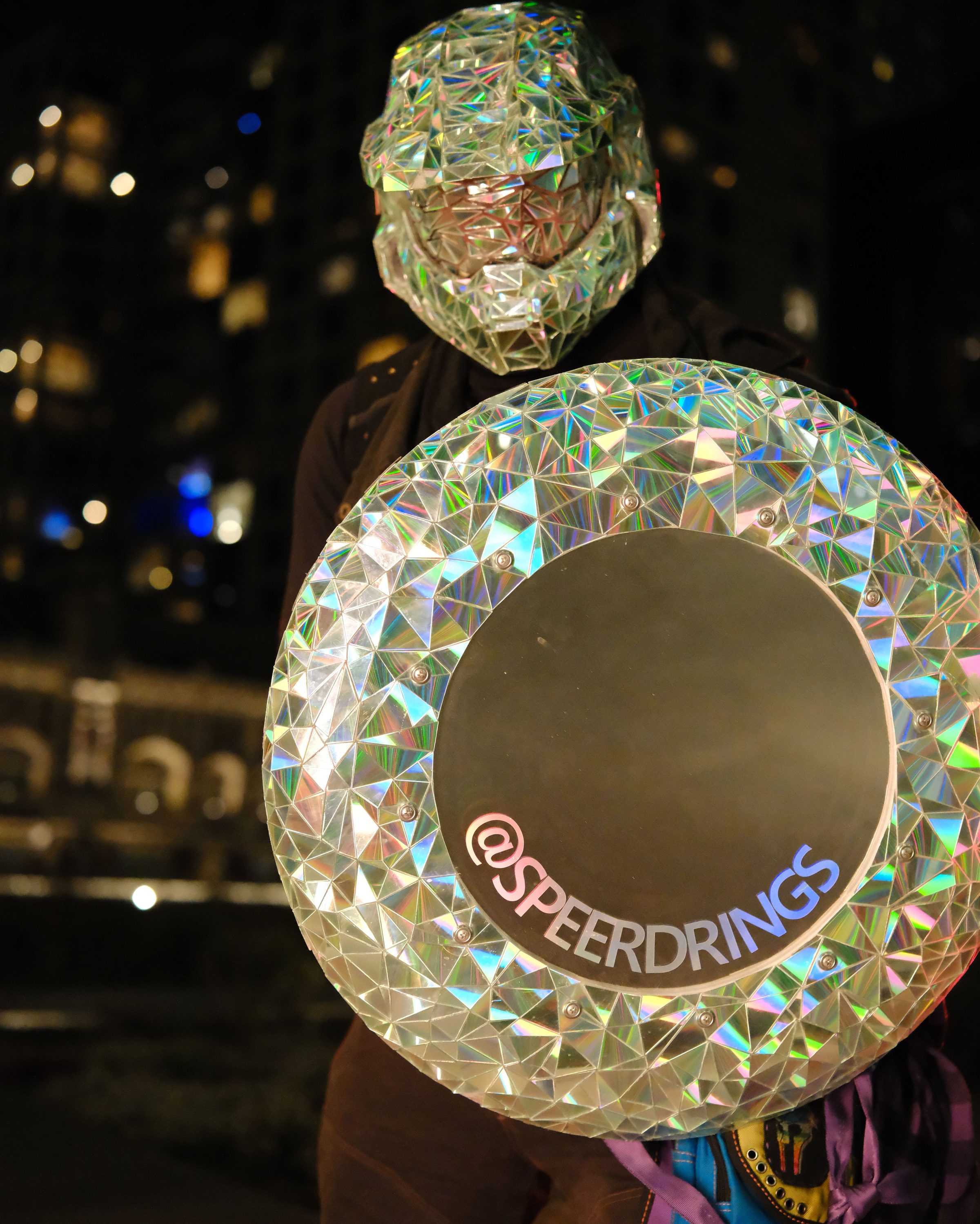 The mirrored helmet and a faceted shield marked @SPEERDRINGS, lit against a city skyline at night.