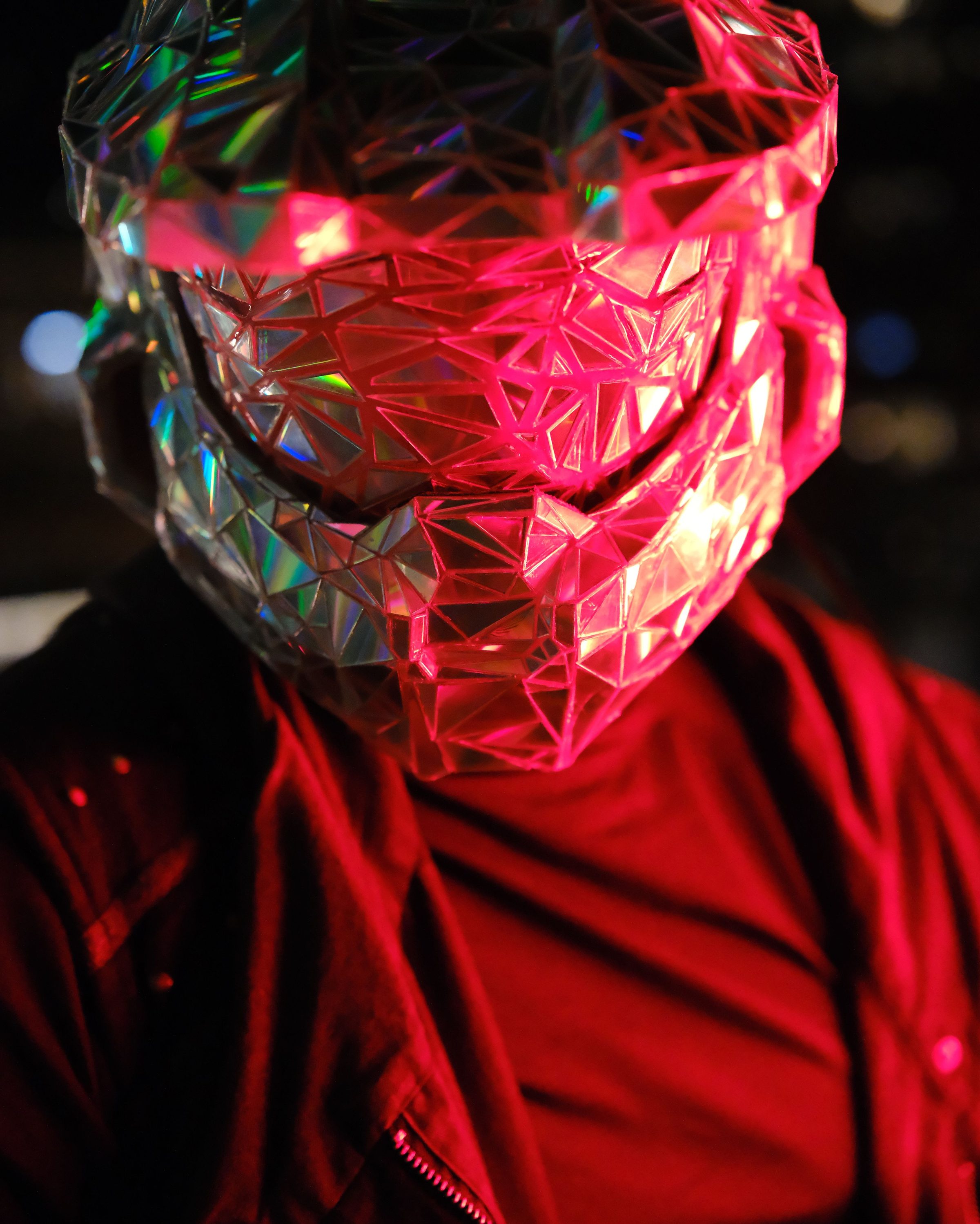 Close portrait of the faceted mirror helmet washed in red light over a red jacket.