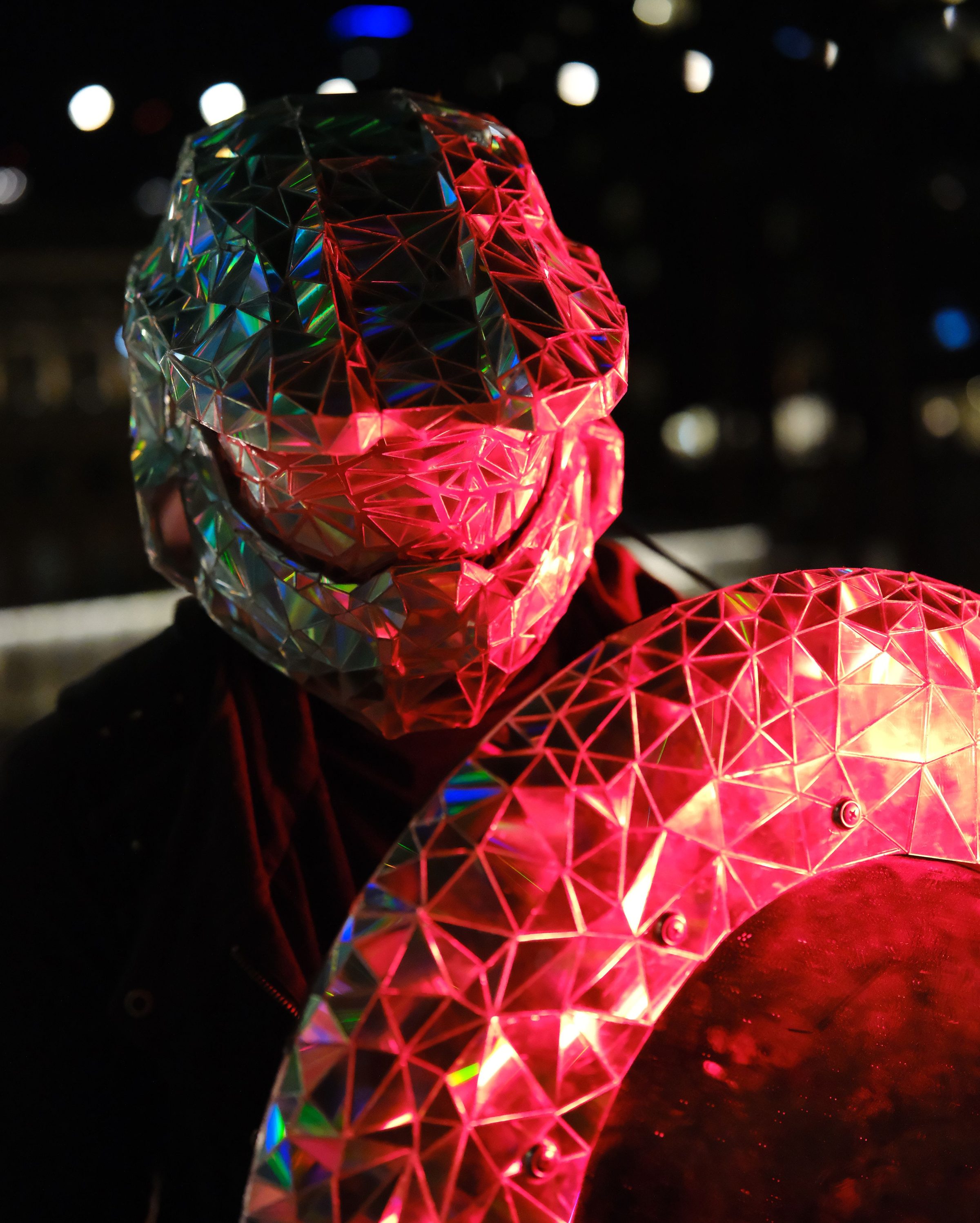The mirror helmet and faceted shield together in red light, city lights blurred behind.