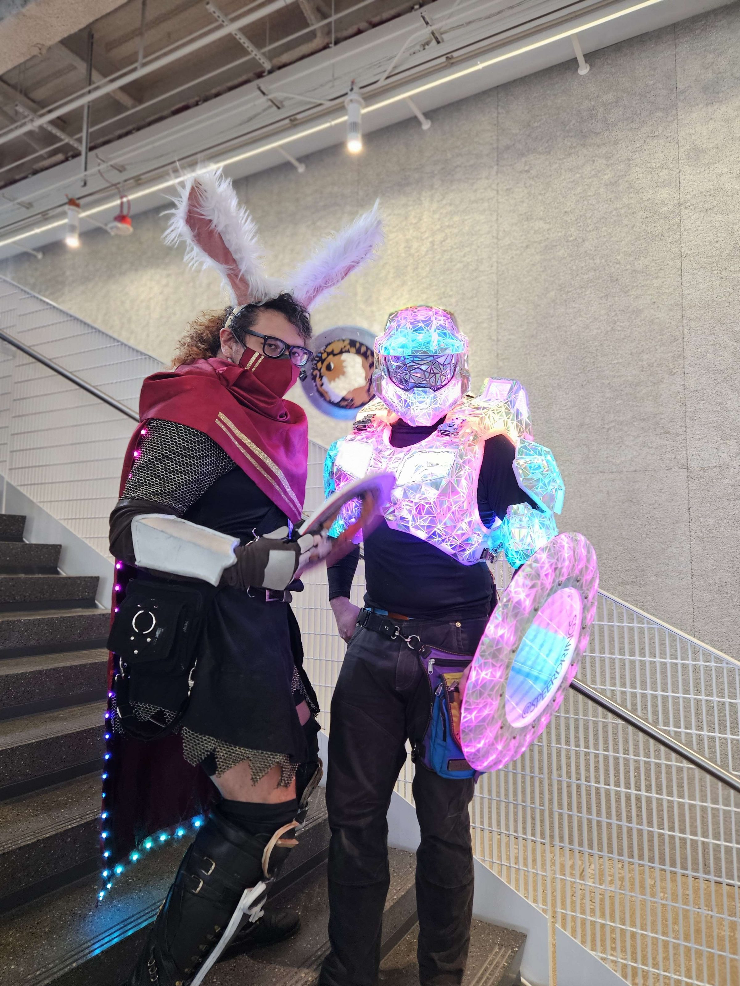 The armour and shield lit pink and cyan in a convention stairwell, beside another costumed guest.