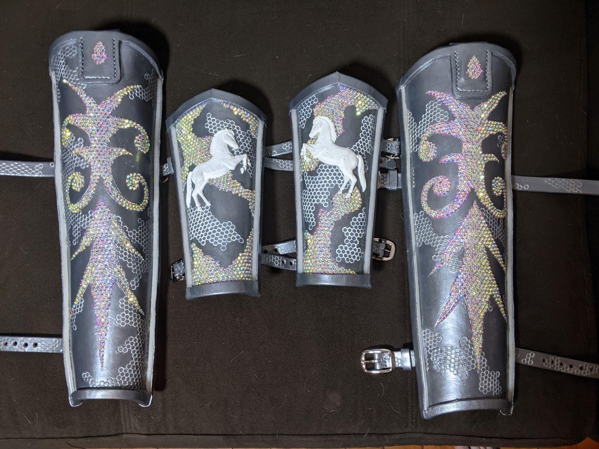 Two greaves and two bracers laid on dark cloth, the greaves in rhinestone filigree and the bracers each carrying a white rearing horse.