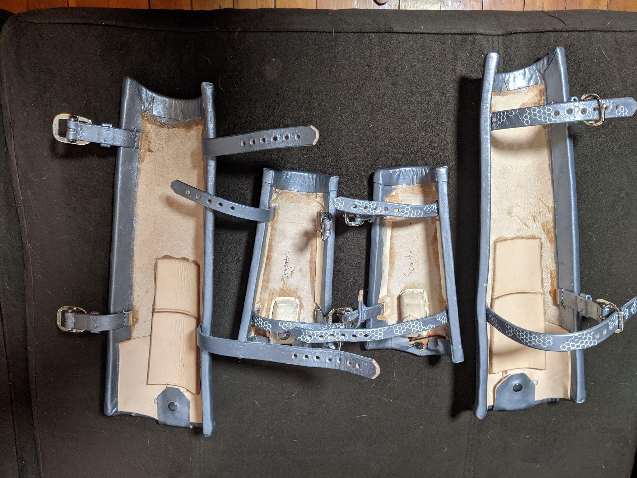 The backs of the greaves and bracers showing raw leather linings, hexagon-punched straps, and buckles.
