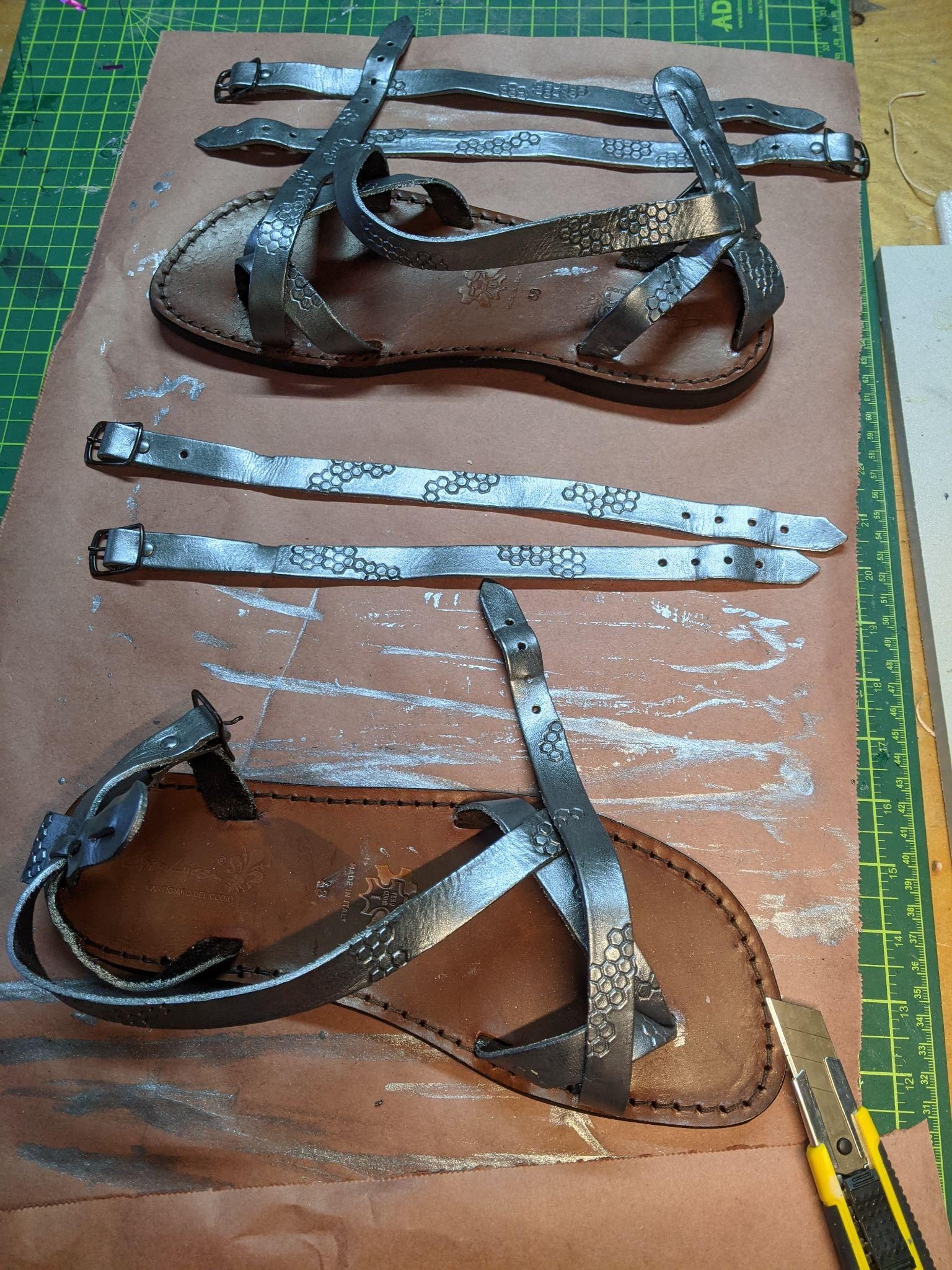 Roman-style leather sandals in progress on a workbench, hexagon-stamped straps painted silver and laid out beside them.