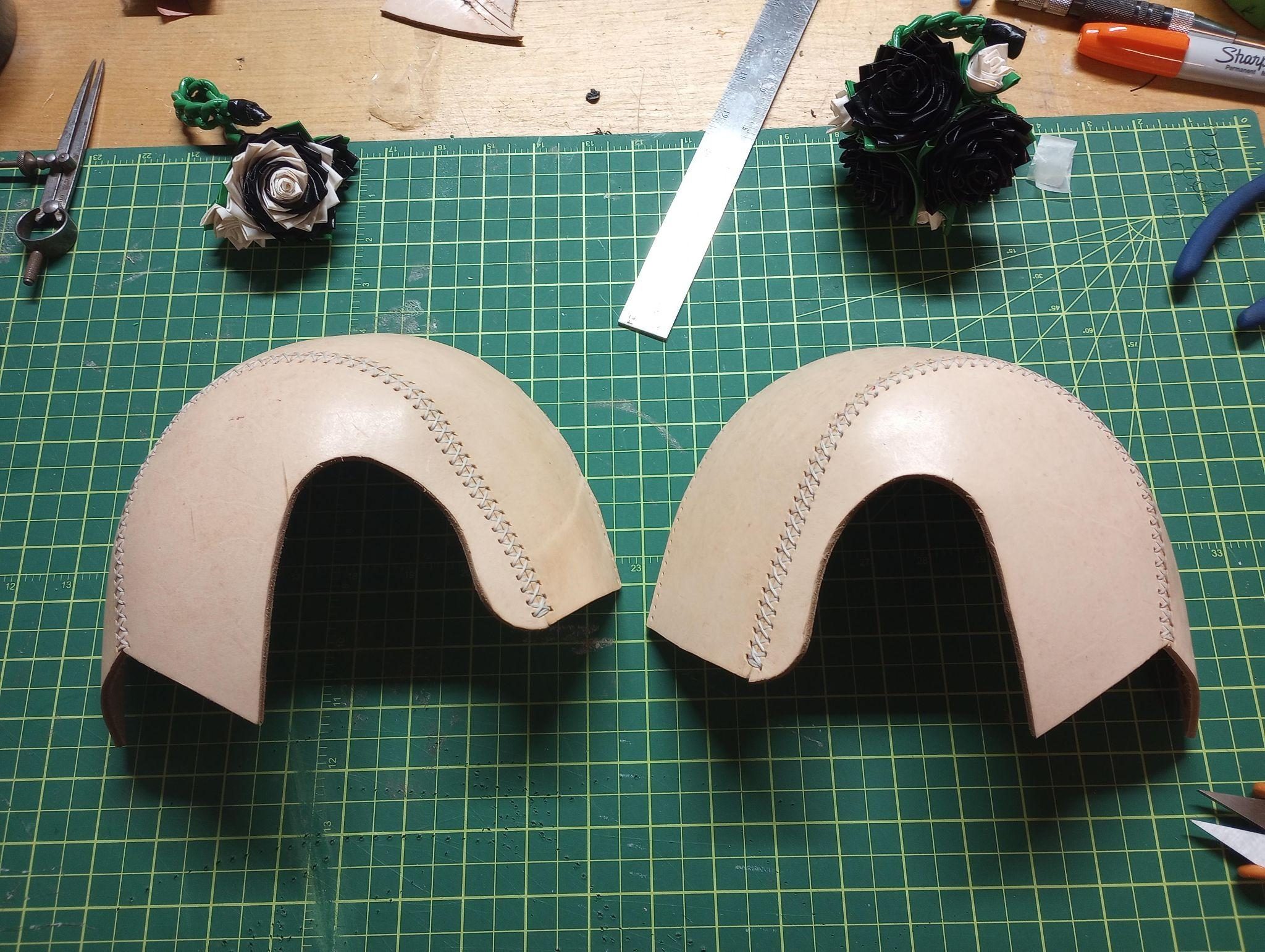 Two hand-stitched veg tan leather helmet halves resting on a green cutting mat beside dividers and a steel rule.
