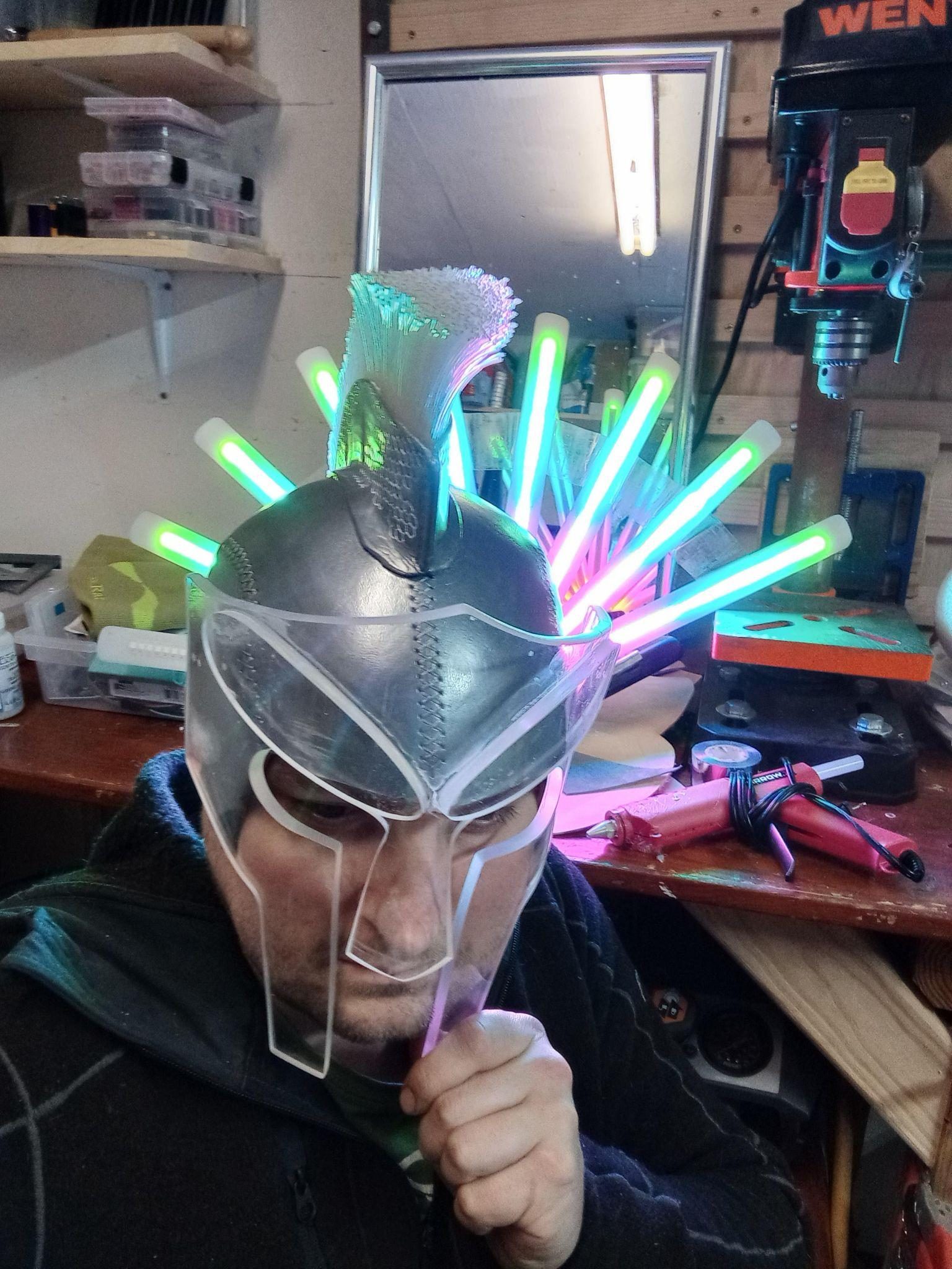 Wearing the painted helmet in the workshop, holding the clear acrylic face plate up against it, LED tubes glowing on the bench behind.