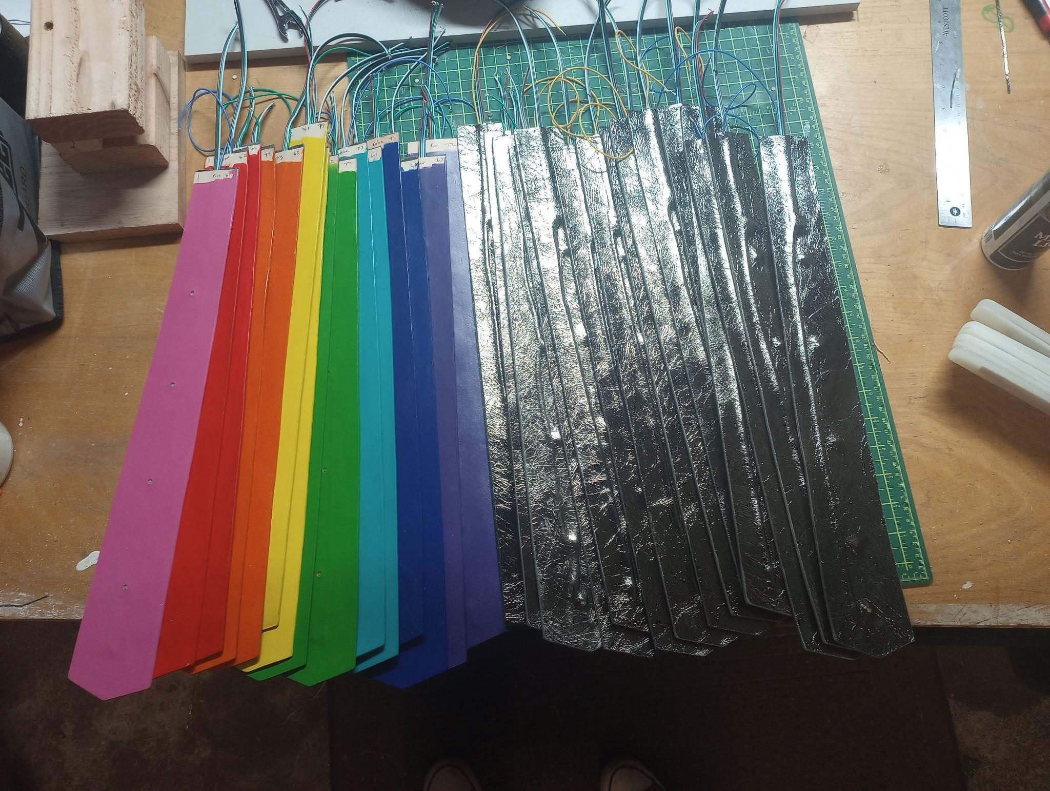 Kilt panels laid out in two sets, one painted in rainbow order and one in silver metallic leather, each trailing its own wires.