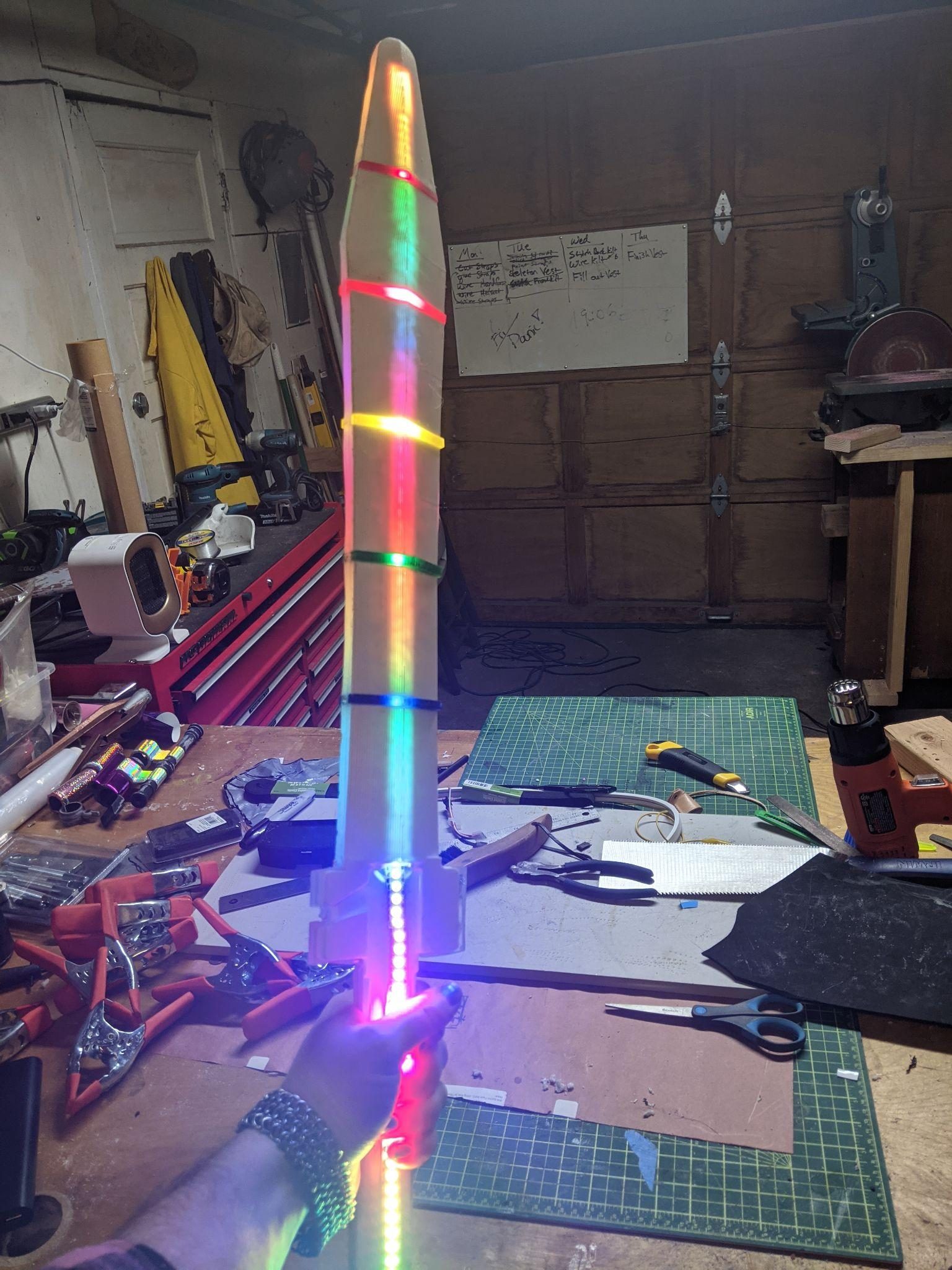 Holding the finished sword upright in the workshop, its blade lit in bands of rainbow colour.