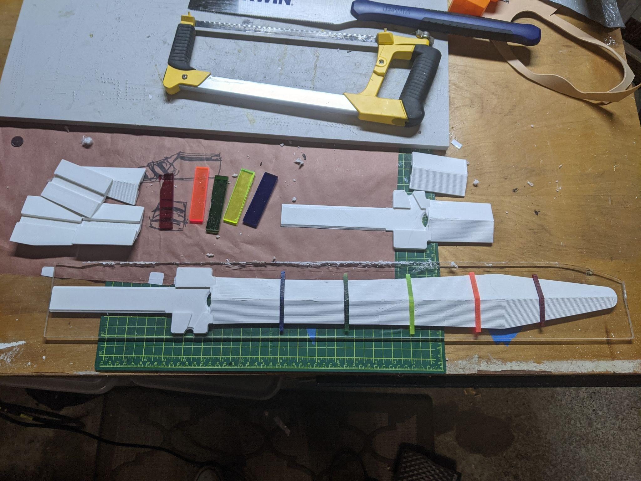 Sword components laid out on the bench: printed white blade panels, the clear acrylic core, offcuts, and a hacksaw.