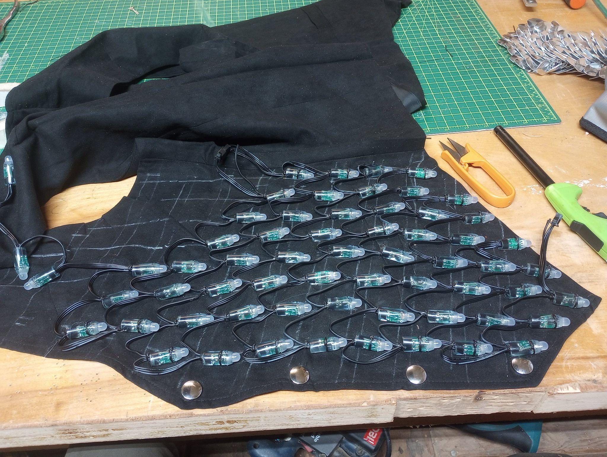 A black vest panel laid flat with rows of LED nodes hand-stitched into a serpentine, wired and marked out in chalk.