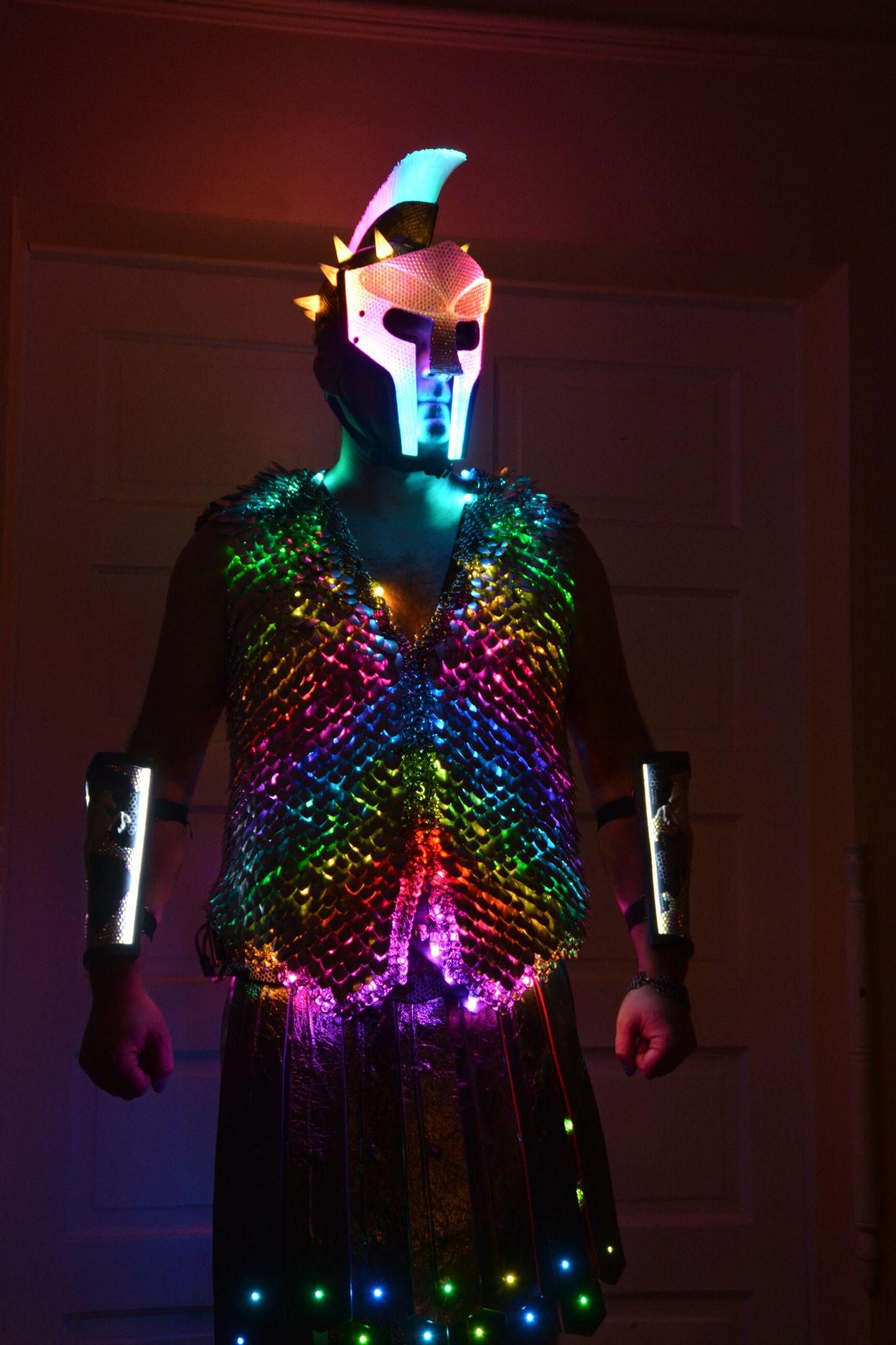 The full costume worn in a darkened room, helmet, scale vest, bracers, and kilt all lit in shifting rainbow colour.