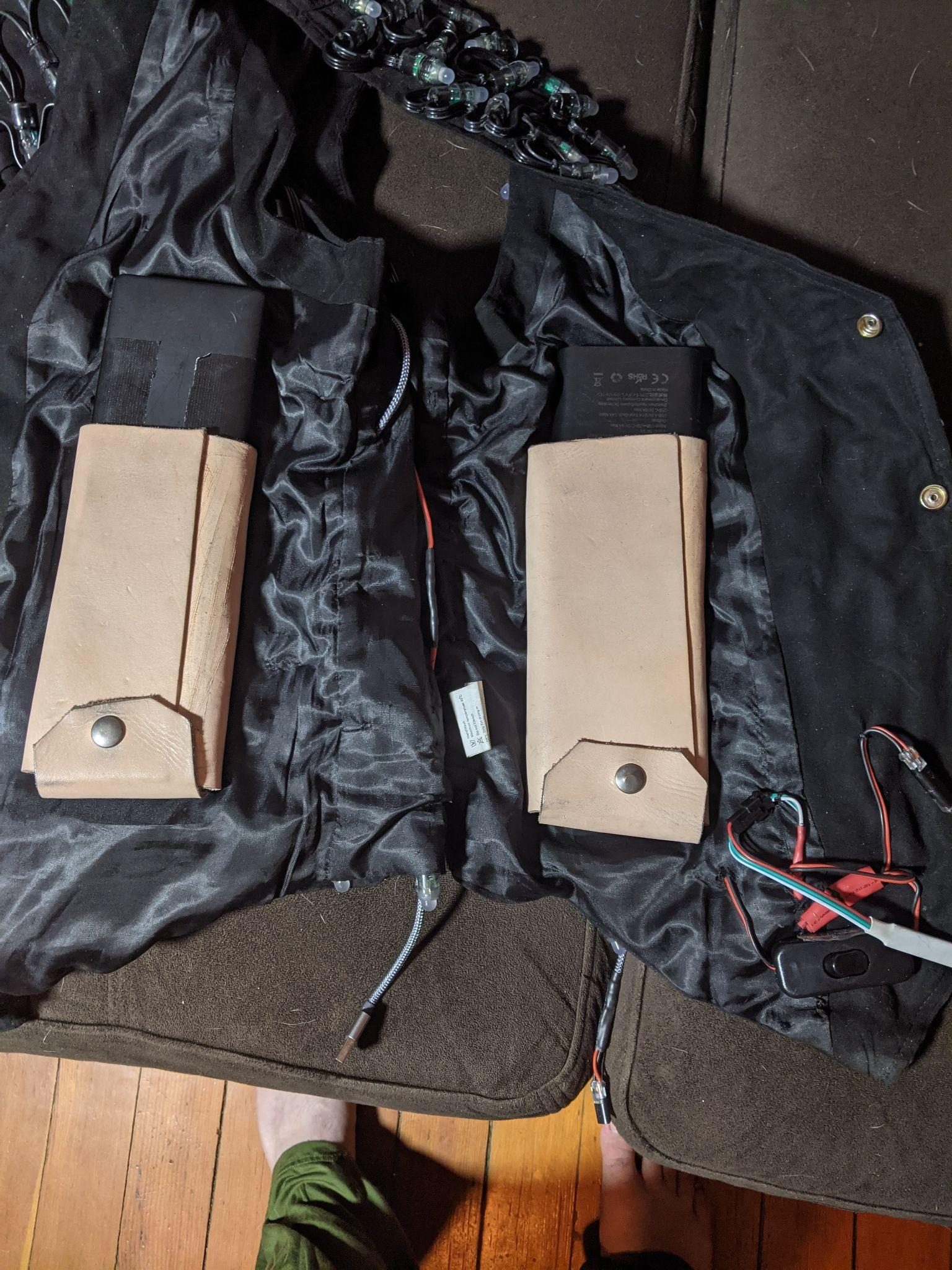 The vest opened flat, showing two leather power-bank pouches with snap flaps, battery packs, wiring, and an inline switch.