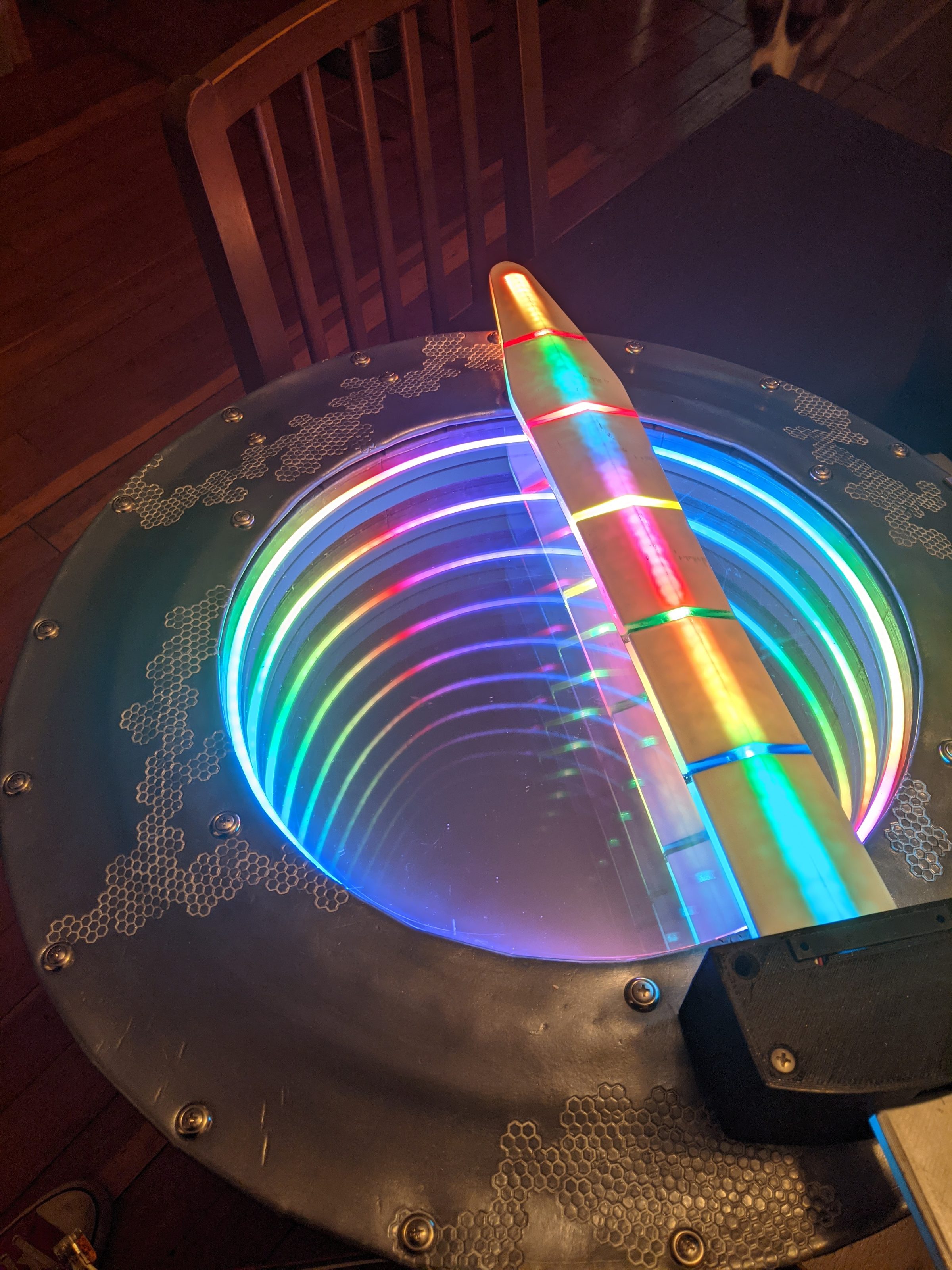 A round metal shield with a rainbow LED spiral and a glowing baton resting across it.