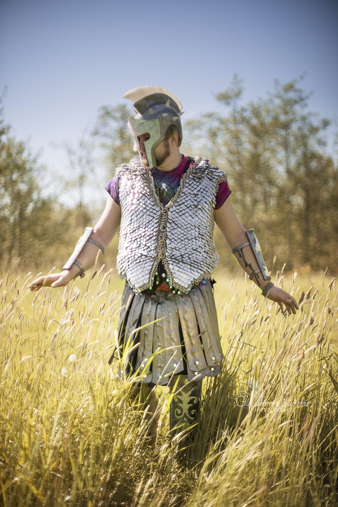 Standing in tall golden grass in daylight, arms out, in the helmet, scale vest, bracers, and kilt.