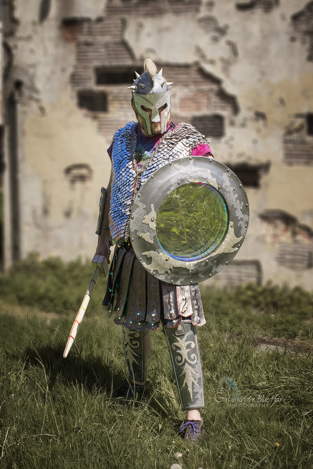 Standing before a ruined wall, shield squared to the camera and the sword held low.