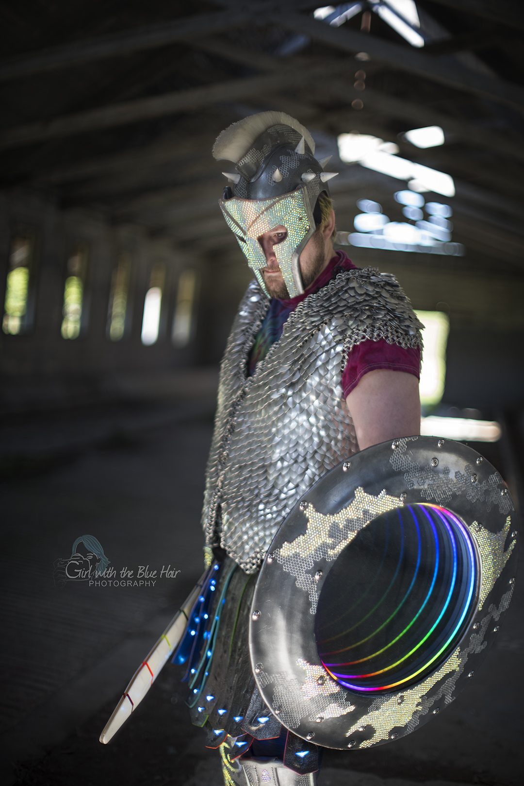 Standing in a covered barn, the shield's mirror lit in rainbow rings and the sword lit at the hip.