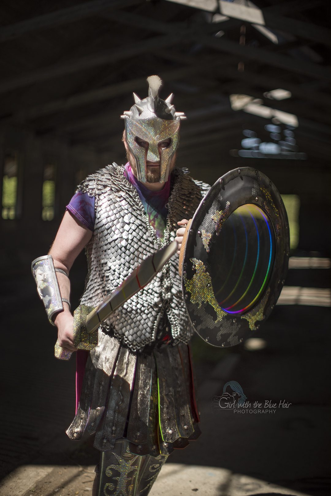 Standing in a barn holding the sword across the body, the shield's mirror lit beside it.