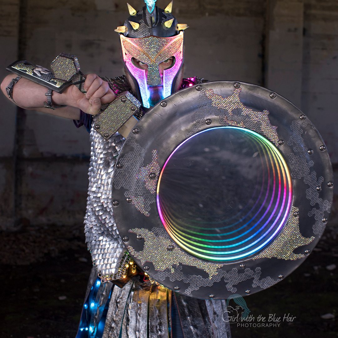 Blade raised beside the rainbow-lit shield, scale maille and glowing visor bright against a bare brick room.
