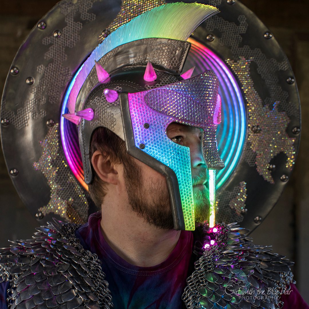 Profile portrait of the helmet, its fibre-optic crest and rainbow visor lit, the shield glowing behind like a halo.