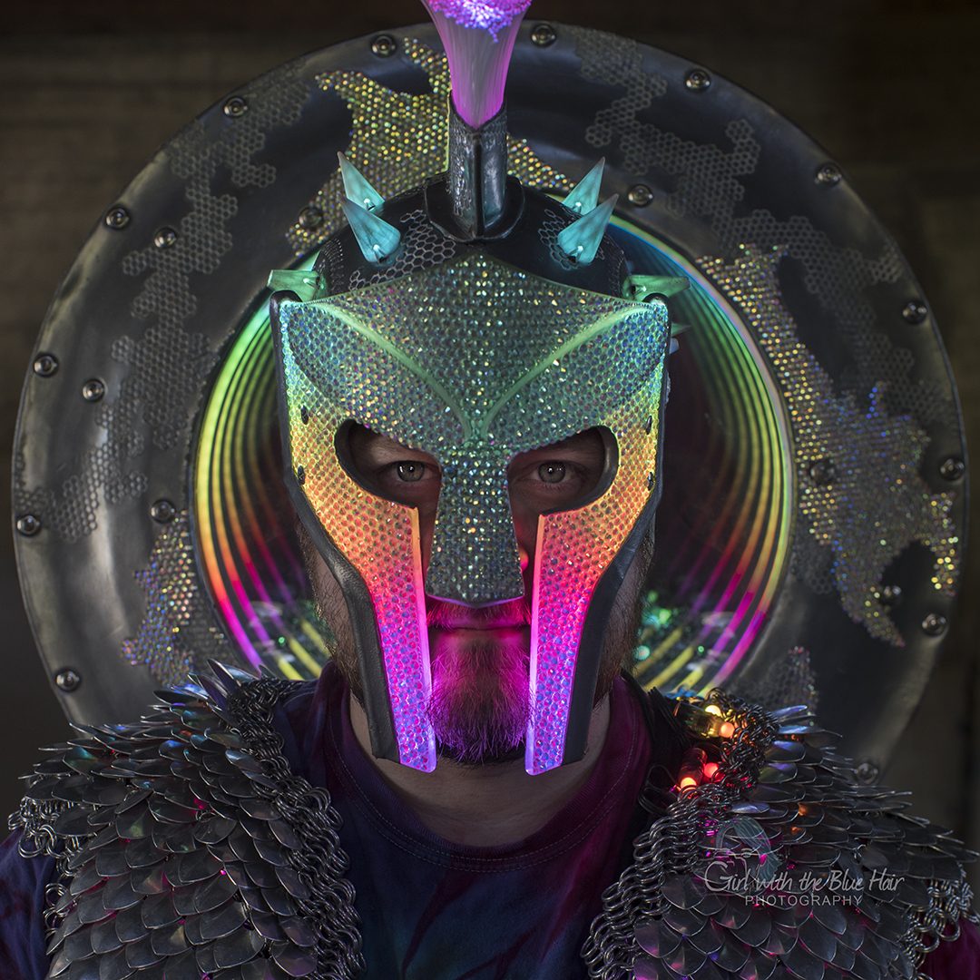Head-on portrait of the rhinestoned helmet glowing green and pink, the rainbow shield haloed behind.