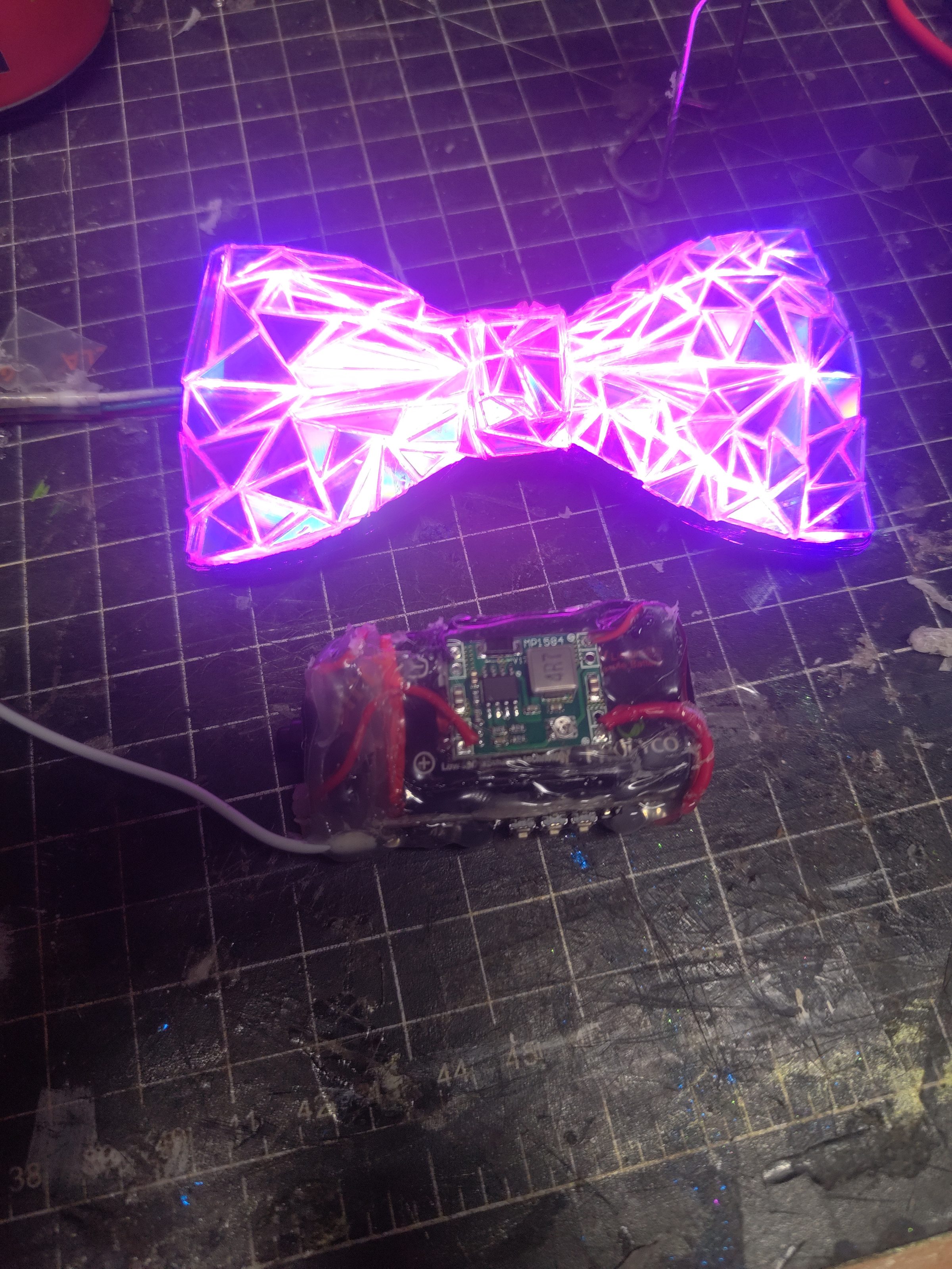 An LED bow tie lit pink, with its controller board on the bench.