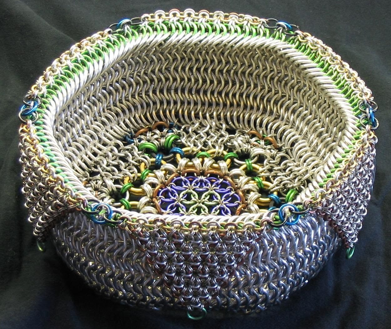 A chainmaille bowl woven from silver rings with green, blue, and gold accents.