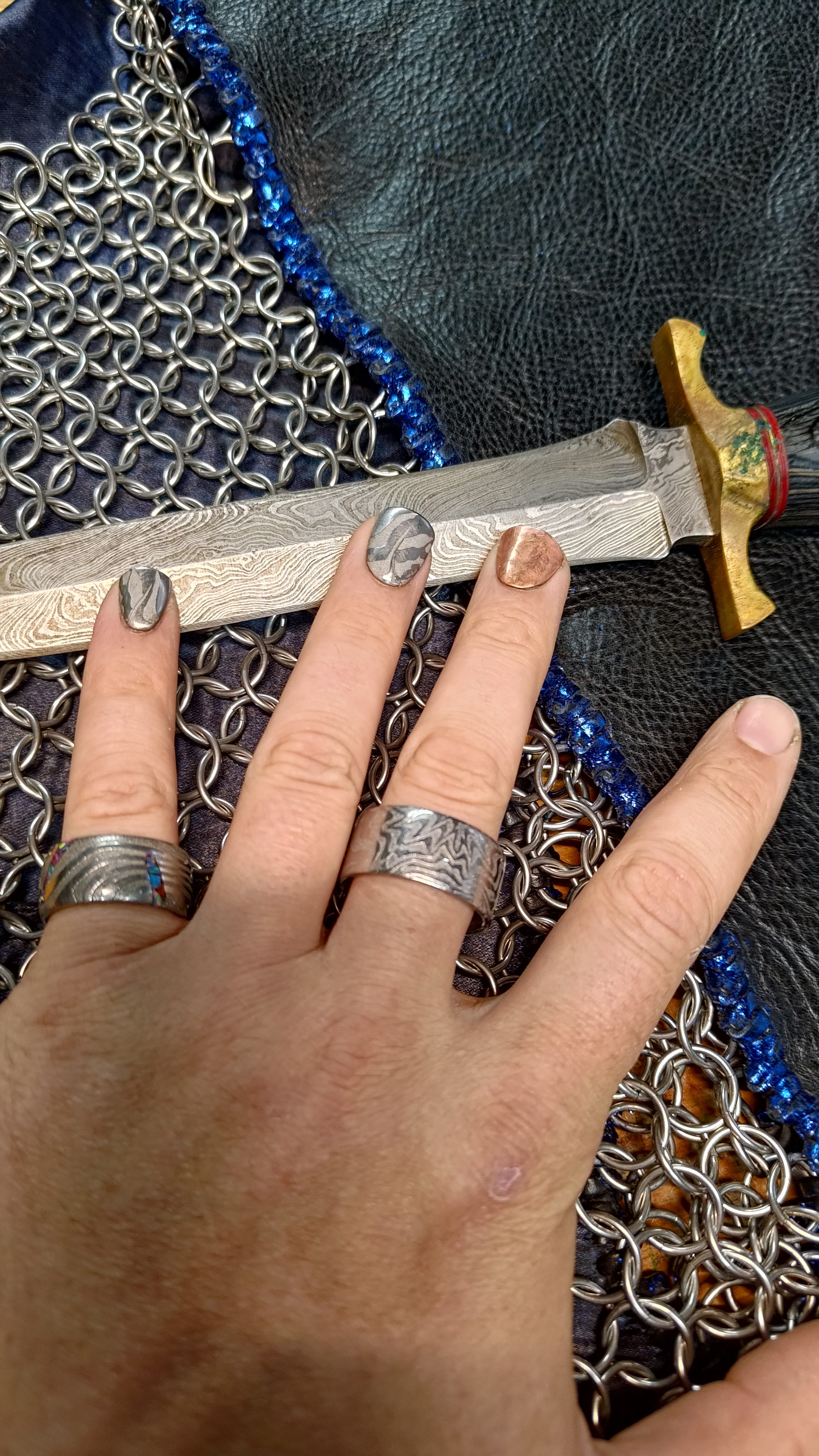A hand wearing two damascus steel rings, resting beside a damascus dagger and chainmaille.