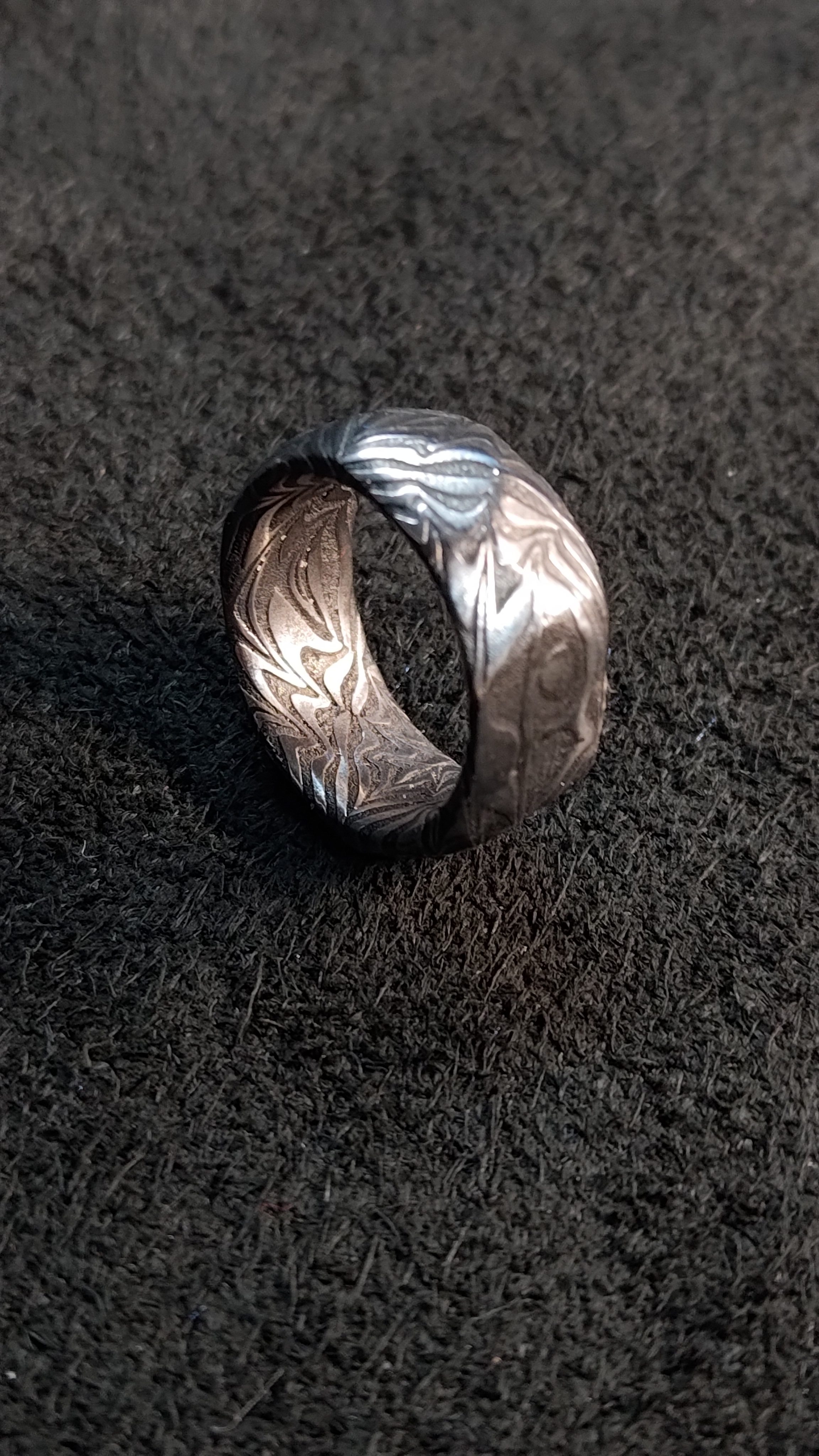 A wide damascus steel ring on a dark background, its pattern running around the band.