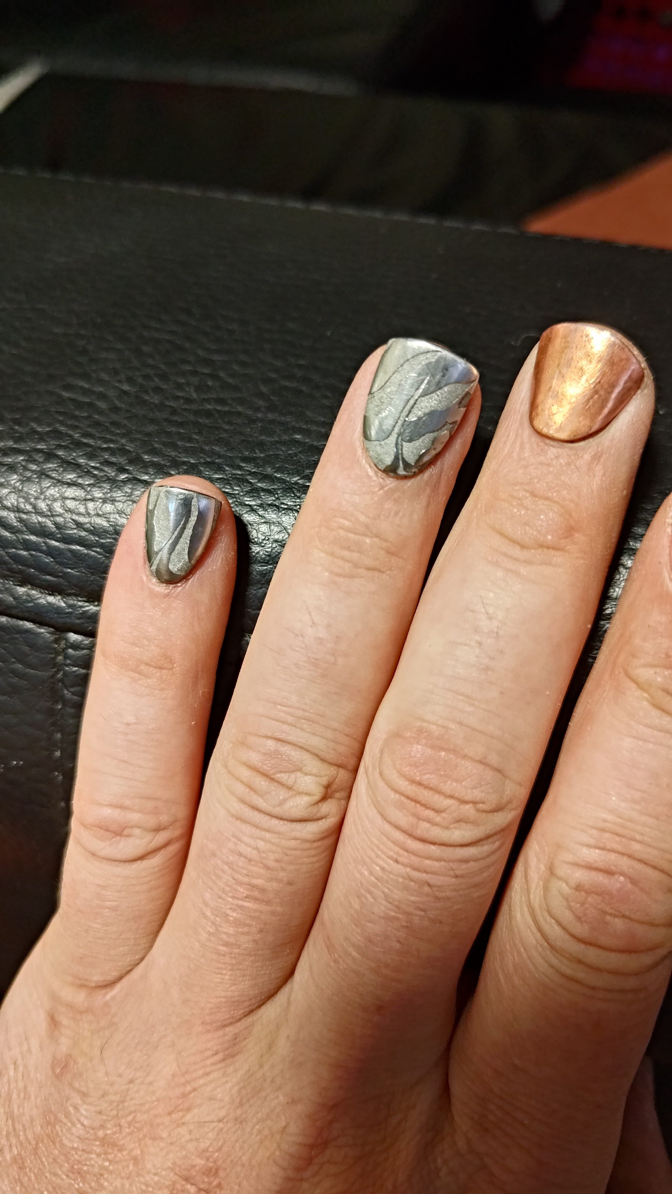 A hand with damascus-patterned and copper press-on nails resting on black leather.
