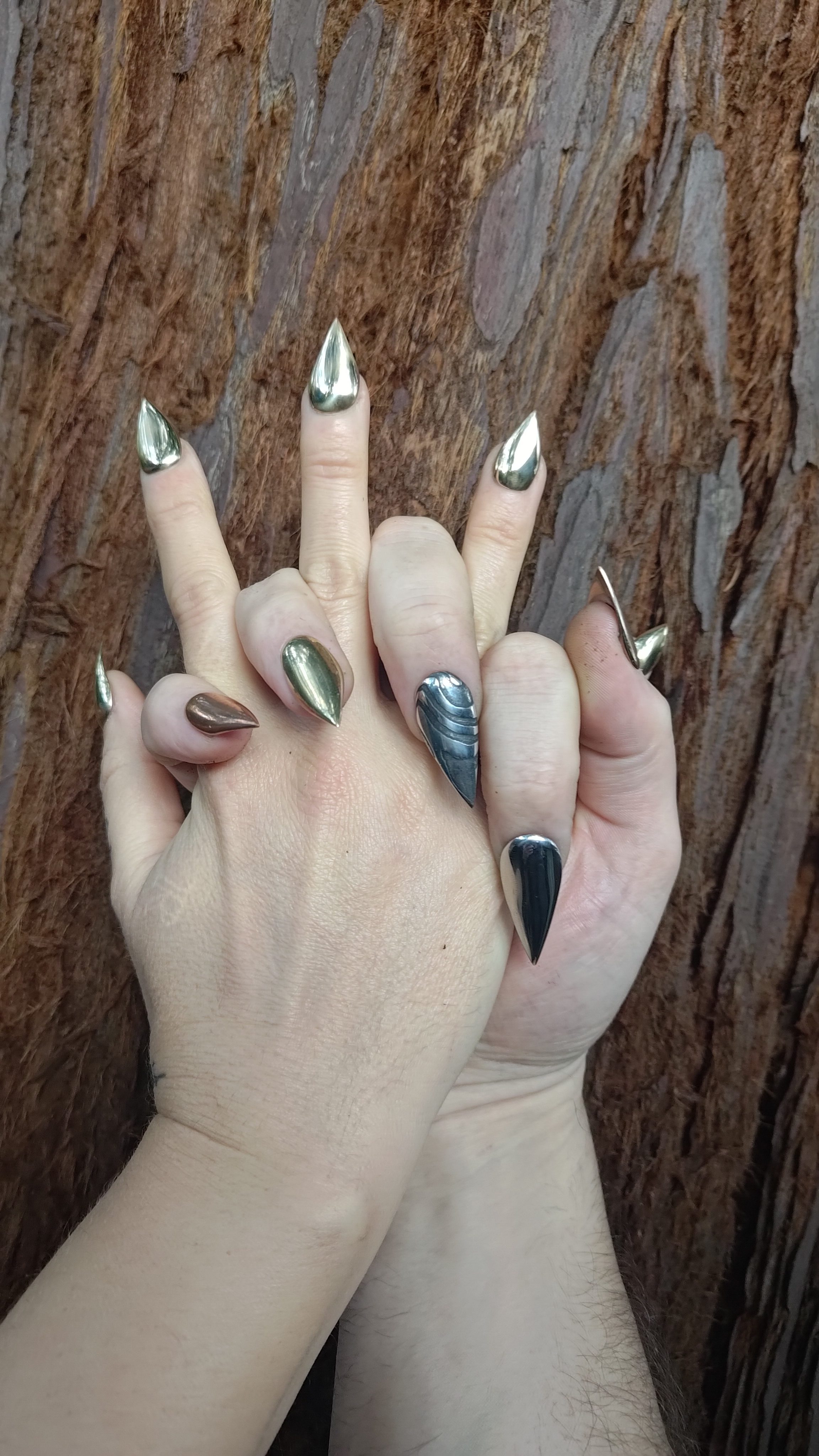 Two hands with long pointed metallic nails against tree bark.